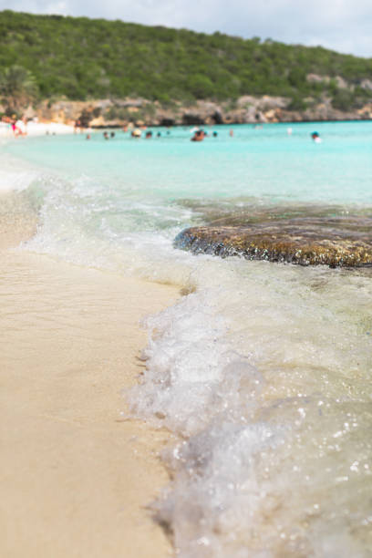 Low-angle perspective of Cas Abao Beach, highlighting a crystal-clear wave gently rolling onto the pristine sands, with the vibrant turquoise waters and tranquil beauty of Curacao in full display.