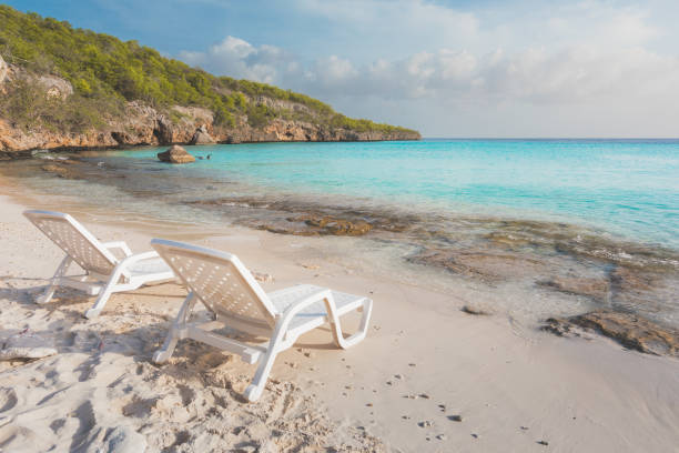 A tranquil scene with two lounge beach chairs set on pristine white sand, as vibrant turquoise waters gently caress the rocky shore. In the distance, reef cliffs draped in lush foliage rise, completing the idyllic backdrop of a perfect Caribbean Sea escape. Captured at sunset in Cas Abao Beach, Curacao.