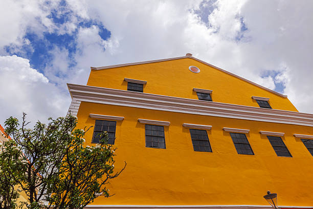 Yellow colonial building with black windows and white trim against blue sky with white clouds. Willemstad. Curacao.
