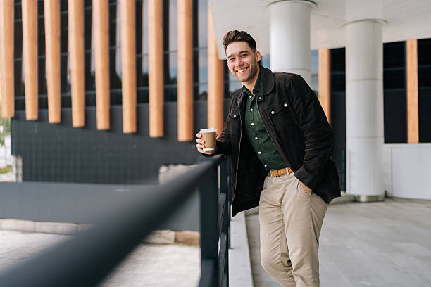 istock Vertical portrait of handsome bearded man holding takeaway coffee cup leaning on railing while standing by modern building, smiling looking at camera. 2206127375