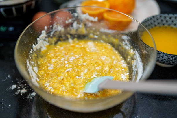 A close-up of a glass bowl filled with freshly mixed orange muffin batter, with flour and zest visible. Halved oranges and fresh juice are in the background, creating a homemade baking scene.