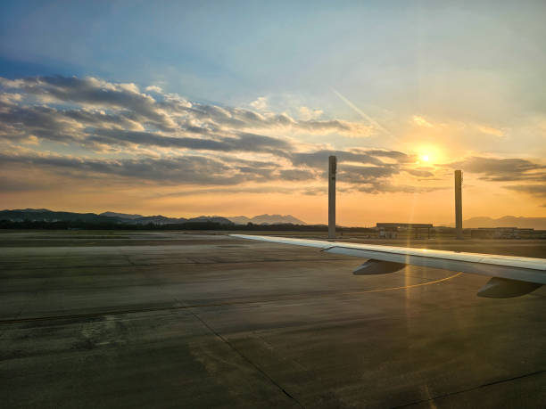 An airplane wing is seen from the cabin window, resting on the runway of an airport during sunset. The tarmac stretches across the foreground while two tall light poles and airport buildings can be seen in the background.