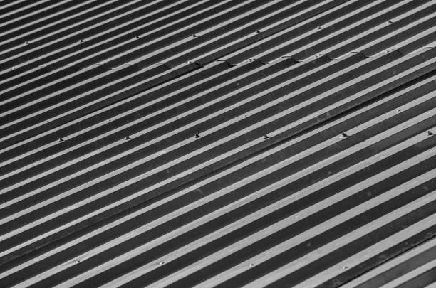 Grey metal roof with bolts. Corrugated metal roof with evenly spaced parallel ridges and visible bolts securing the panels. The surface appears to be a dark grey, suggesting durability and weather resistance. The bolts are in neat rows along the ridges, indicating structural reinforcement. The repetitive pattern and texture create a sense of uniformity and industrial design, commonly used in commercial or warehouse buildings for efficient water drainage and protection. The material likely provides long-lasting coverage with minimal maintenance.