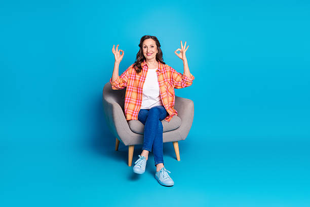 istock Smiling middle-aged woman in stylish checkered shirt doing hand gestures while seated on chair against vibrant blue background 2206179433