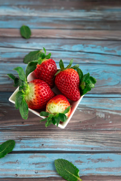 Fresh strawberries in square bowl on rustic blue wood background, decorated with mint leaves. Copy space.