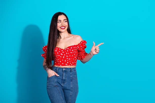 istock Smiling young woman in a red off-shoulder top posing against a vibrant blue backdrop with a hand gesture of pointing 2206181632