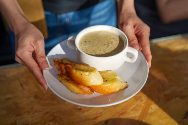 A warm bowl of creamy mushroom soup paired with crispy garlic bread, served on a white plate. A person is holding the plate, ready to enjoy this comforting appetizer.