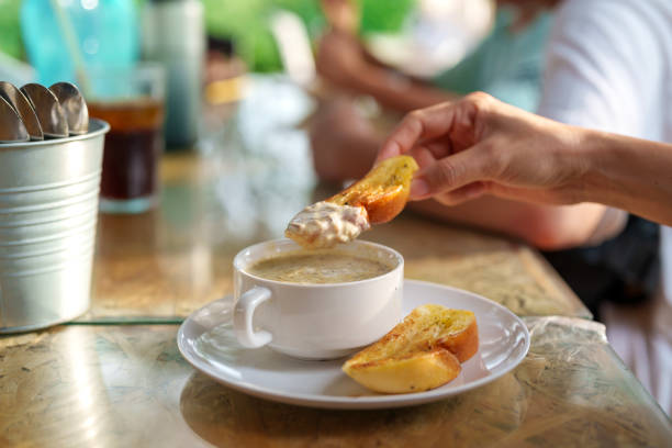 A person dipping a crispy slice of garlic bread into a cup of creamy mushroom soup at a casual dining setting. A comforting and delicious appetizer enjoyed in a relaxed environment.