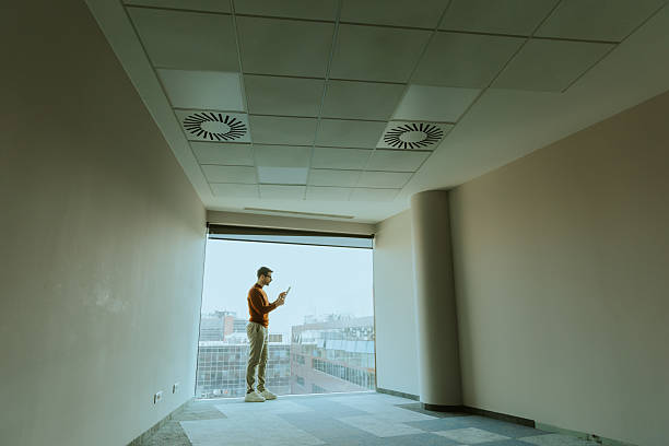istock Man standing in a spacious room with cityscape view, engaged with his mobile device 2206185990