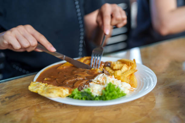 A freshly served plate of crispy chicken chop covered in rich mushroom gravy, accompanied by crinkle-cut fries, coleslaw, lettuce, and an omelet. A person is cutting the meal with a fork and knife at a casual dining restaurant.