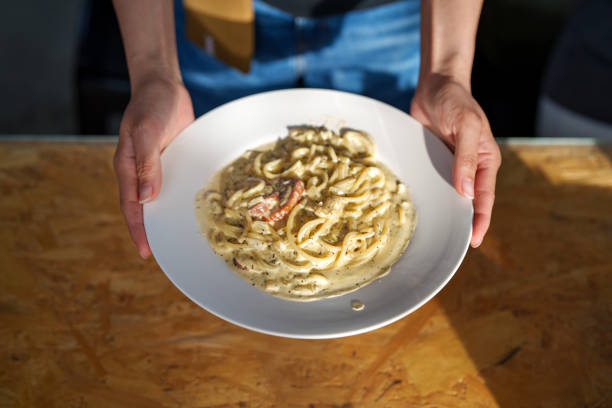 A freshly prepared plate of creamy carbonara pasta with a rich sauce, garnished with herbs and crispy bacon. A person is holding the plate, presenting the delicious meal on a rustic wooden table.