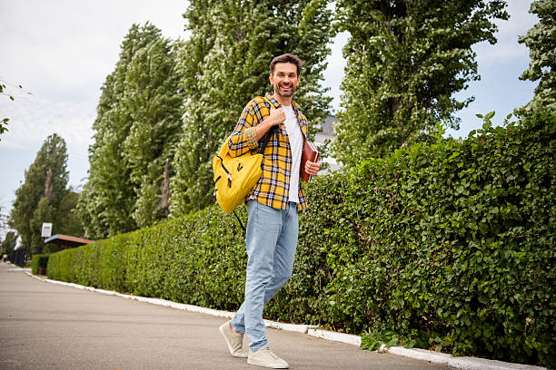 A handsome young man in a checkered shirt and jeans with a yellow backpack, walking along a green hedge in daylight, smiling.