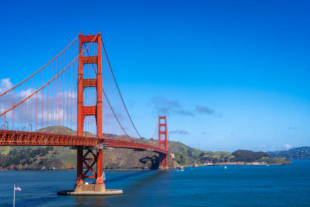 The Golden Gate Bridge stretches across the bay, its red towers contrasting against the blue sky and water, with green hills in the background