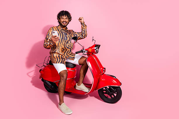 istock Young stylish man on pink background posing on a red scooter while holding a smartphone and showing a triumphant 2206190529