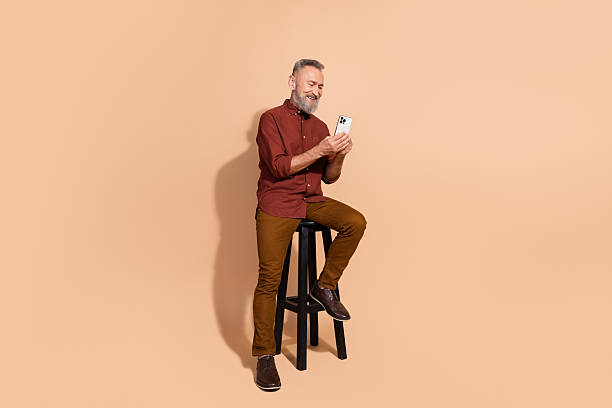 istock Smiling senior man with gray beard browsing smartphone while sitting stylishly on stool against beige background in studio setting 2206191255