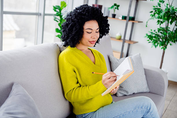 istock Young stylish woman writing in her notebook while sitting on a cozy sofa in a modernly designed daylight-lit living room. 2206195460