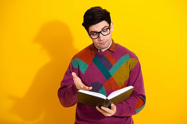 istock Stylish man wearing glasses reading a book while gesturing thoughtfully against a vibrant yellow background in a casual retro sweater 2206197779