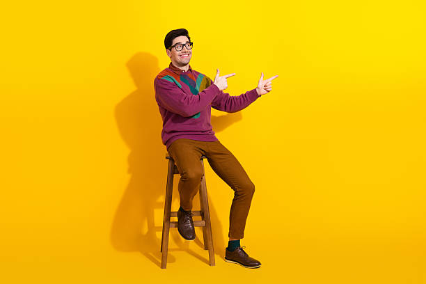 istock Cheerful stylish young man on a stool making pointing gestures against a vibrant yellow background 2206197892