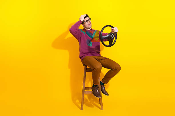 istock Young man posing on a stool with hand on his head while holding a steering wheel against a vibrant yellow background 2206197899