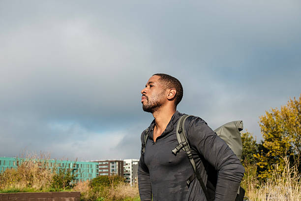 Athletic man with a backpack explores nature near a city, gazing up at the cloudy sky, symbolizing strength and perseverance while seeking adventure and challenges