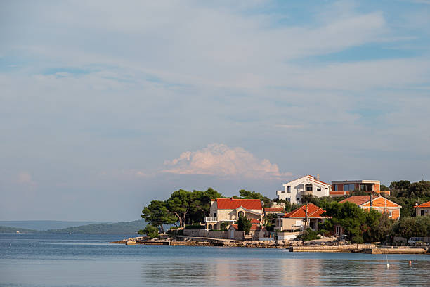 Charming Mediterranean village with red-tiled roofs and lush greenery nestled on a peaceful coastline under a bright, partly cloudy sky.