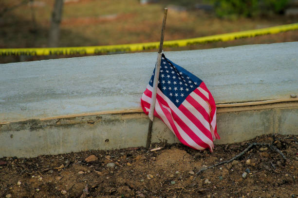 Small tattered flag of the United States placed in dirt with a strip of yellow caution tape visible in the background.