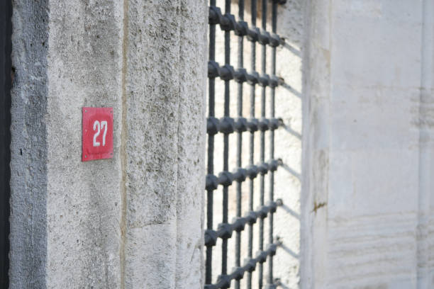 Closeup of a jail cell door marked 27, highlighting sturdy metal bars and harsh concrete