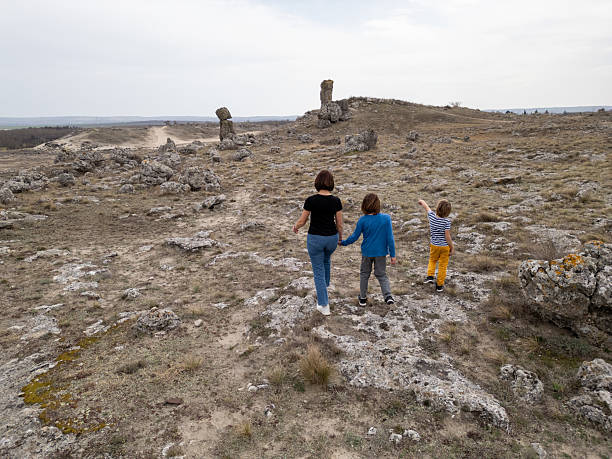 istock A mother and two child exploring a rocky outdoor landscape. The terrain is rugged, with sparse vegetation and distant hills in the background 2206232202