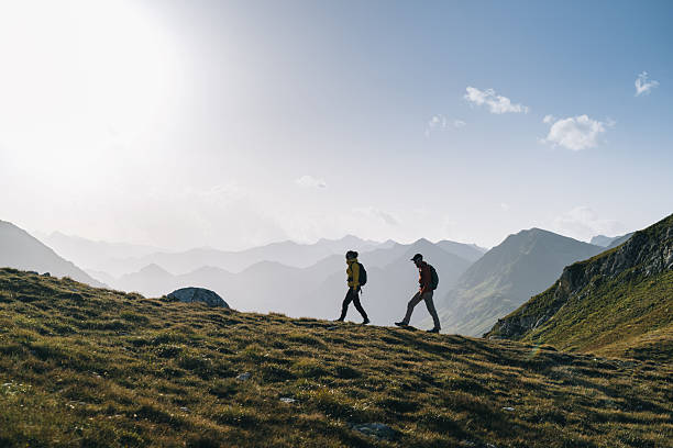 Couple hike across mountain meadow at sunrise, side view, Ticino, Swiss Alps