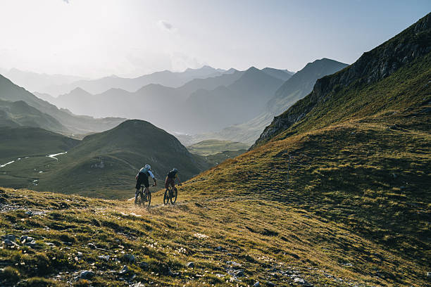 Mountain biking couple down mountain trail, valley and mountain ranges illuminated by morning sunlight, Ticino, Swiss Alps