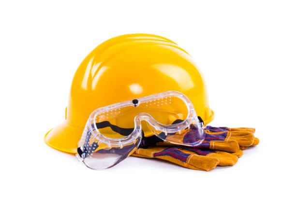 A yellow hard hat rests alongside a pair of work gloves and clear protective goggles, all intended for safe practices in construction or industrial activities. The arrangement emphasizes essential safety equipment commonly used on job sites.