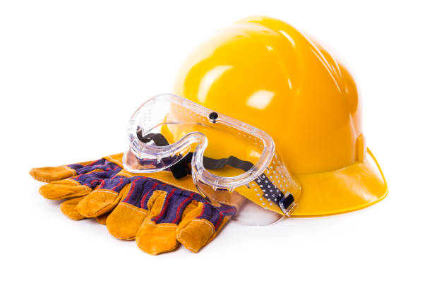 A yellow hard hat sits alongside protective goggles and glazed work gloves on a clean white backdrop, highlighting crucial safety equipment used in construction and industrial settings to ensure worker safety during tasks.
