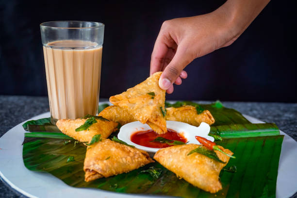 Image of hand dipping samosa into tomato sauce and ketchup with chai tea, banana leaf in sri lankan fast food style