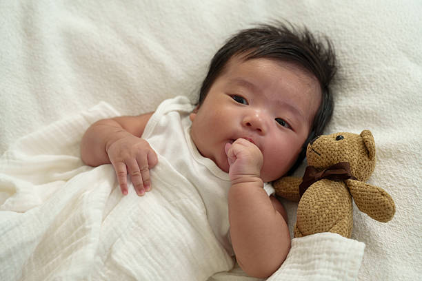 istock Asian newborn baby lying on a soft white blanket, gently sucking thumb while cuddling a small teddy bear. Representation of warmth, comfort, and security, symbolizing early childhood development. 2206250344