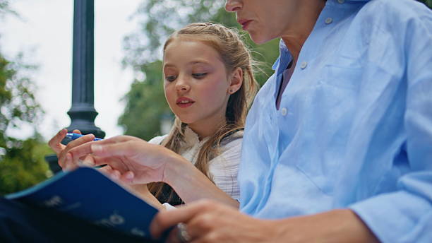 Closeup schoolgirl talking mom at park. Cute small kid writing copybook studying with female parent together at wooden bench. Positive curious toddler looking book asking questions. Family concept
