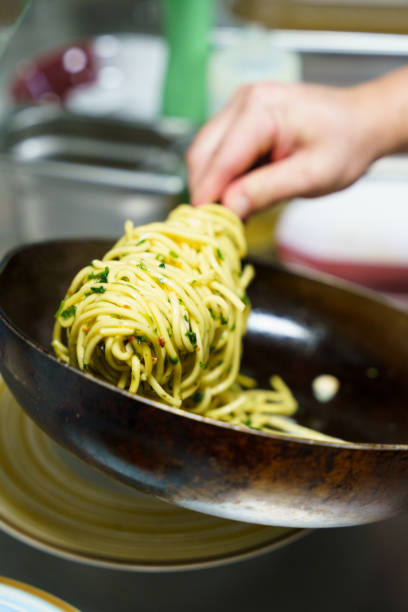 A chef carefully plates a serving of spaghetti aglio e olio using tongs in a professional kitchen. The pasta is coated in olive oil, garlic, parsley, and red pepper flakes.