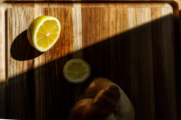 A fresh yellow lemon cut in half sits on a wooden cutting board. Dramatic sunlight creates a strong diagonal shadow across the wooden surface, highlighting the citrus fruit's texture and bright color. The rustic wood grain of the board provides contrast to the vibrant fruit. The composition shows selective focus on the lemon slice with its visible segments and pith.