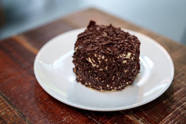 Close-up of a small round chocolate cake covered in dark chocolate shavings served on a white plate in a cozy indoor setting.