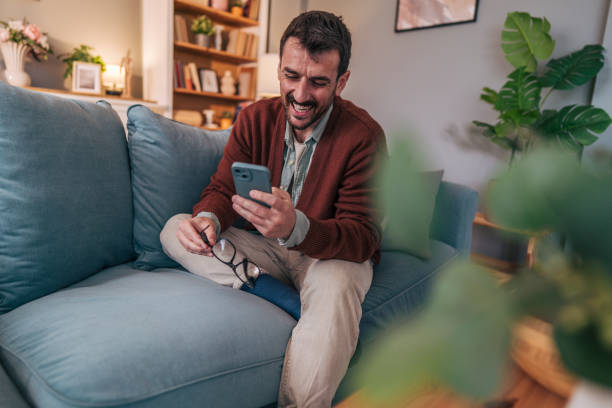 istock Happy man using smartphone and holding eyeglasses while relaxing on sofa at home 2206338926