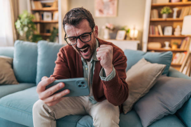 istock Excited man celebrating success while playing mobile game on sofa 2206339491