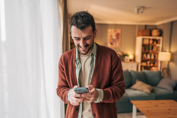 istock Smiling man using smartphone at home standing near window 2206339750