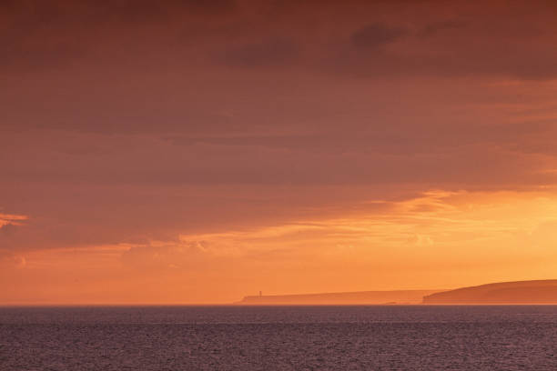 seascape view in County Wexford, Ireland, featuring the vast expanse of the ocean and a distant coastline. The setting sun casts a warm, orange glow over the water and sky, creating a dramatic and picturesque scene. The distant coastline is silhouetted against the horizon, adding depth to the image. The sky is filled with clouds, enhancing the sunset's colorful effect. The image conveys a sense of tranquility and the natural beauty of the Irish coastline at dusk.