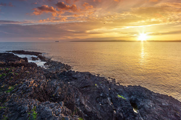 sunset view of the coastline in County Wexford, Ireland, featuring a rocky shore and the vast expanse of the ocean. The setting sun casts a warm, golden light over the water, creating a serene and picturesque scene