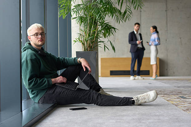 istock A casual young professional sitting by a large window in a modern office, smiling while working on a laptop, with colleagues interacting in the blurred background. 2206357743