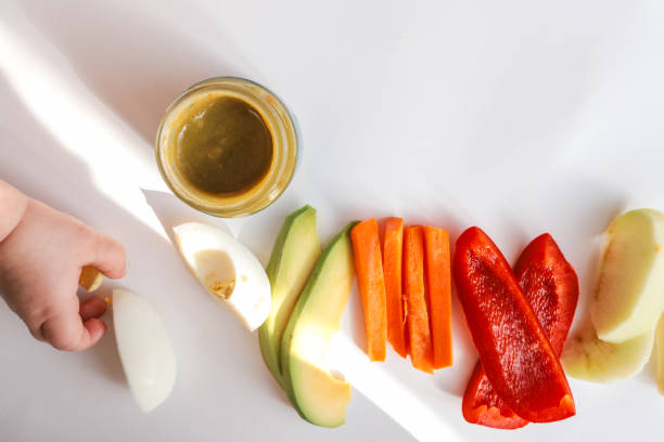 istock A child's hands reaching for freshly chopped vegetables on a white surface. Includes diced avocado, carrot, apple slices, bell pepper, and tomato pieces. Minimalist food composition. 2206357970