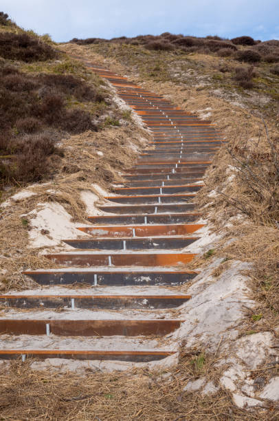 Rusty metal staircase climbing a sandy hillside covered in dry grass, creating a contrast between nature and man-made structures