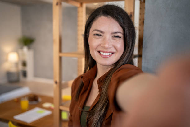 istock Young woman taking a selfie in home office with arm extended 2206367334