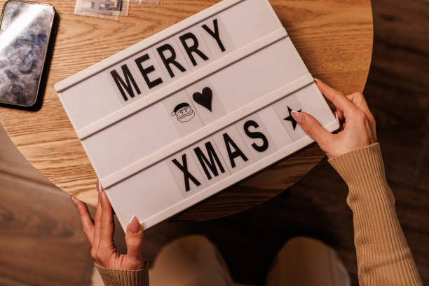 Hands holding a festive lightbox sign displaying 'MERRY XMAS' above a wooden table, embodying the warm and joyful holiday season. Cozy indoor atmosphere with celebratory Christmas elements and a smartphone nearby.