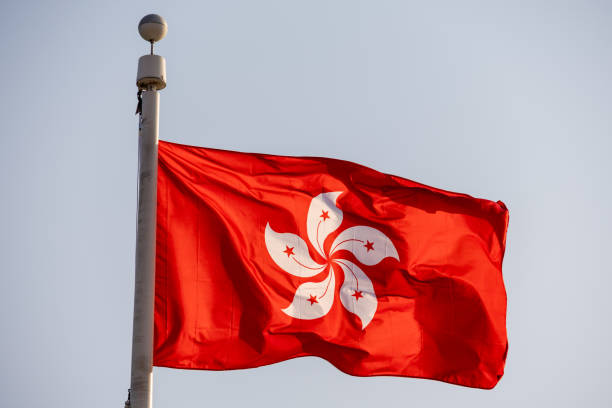 The flag of Hong Kong is waving in the wind, featuring a white, five-petal Bauhinia flower with red stars on a vibrant red background. The motion highlights the flag's smooth fabric texture and symbolic design.