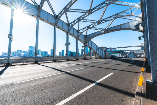 Empty highway under metal bridge leading to tokyo skyline with skyscrapers and bridge on sunny day with lens flare